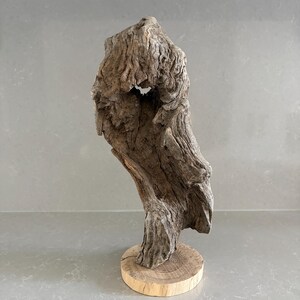 May include: A piece of weathered, textured driftwood with a natural hole near the top, mounted vertically on a circular, light-colored wooden base. The wood displays deep grooves, ridges, and a gray-brown color throughout its organic form.