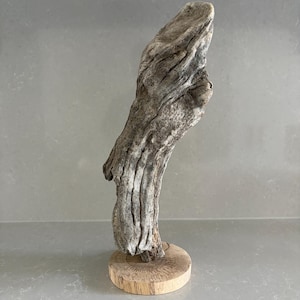 May include: A piece of weathered, grey driftwood with a textured, gnarled surface standing upright. The driftwood is securely mounted onto a circular, light-colored wooden base that features visible grain patterns and a smooth, flat top surface.