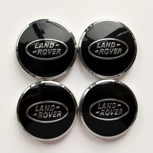 May include: Four black Land Rover wheel centre caps with a silver trim. Each cap features the Land Rover logo in silver lettering on a black background. The caps are circular and have a glossy finish.