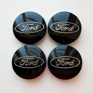 May include: Four black Ford hubcaps with a glossy finish. Each hubcap features the Ford logo in silver script within a silver oval border. The hubcaps are arranged in a square, highlighting their design and finish.