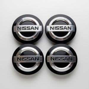 May include: Four black and silver Nissan hubcaps. Each hubcap has a black center with a silver ring around the edge. The word "NISSAN" is displayed in silver lettering across the center of each hubcap.
