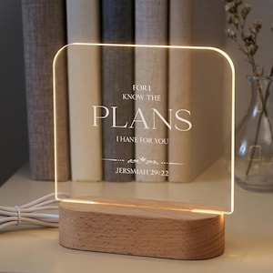 May include: A lighted acrylic sign with the text "FOR I KNOW THE PLANS I HAVE FOR YOU JERMSIAH 29:22" sits on a wooden base. The sign is illuminated with a warm glow. The background includes books and a vase with dried flowers.