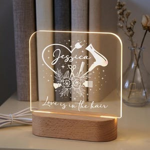 May include: A clear acrylic LED lamp with a wooden base. The lamp features a heart design with the name "Jessica", a hairdryer, scissors, and the text "Love is in the hair". The lamp is on a white surface.
