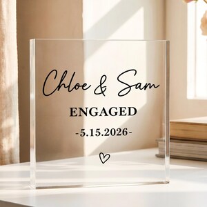 Può includere: Un blocco quadrato in acrilico trasparente con testo stampato in nero che recita Chloe & Sam, ENGAGED e la data 5.15.2026, rifinito con un piccolo simbolo a forma di cuore disegnato a mano e centrato sul fondo dell'oggetto decorativo.
