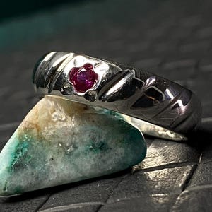 May include: A silver ring with a small, round, red gemstone set in the center. The ring has a textured design with diagonal lines and is resting on a small, multi-colored stone with green and tan hues.