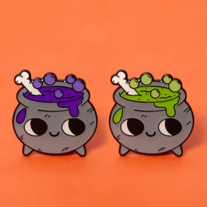 May include: Two enamel pins shaped like cauldrons with cute faces. One cauldron is filled with purple potion and the other with green potion. Both have a bone sticking out of the top.