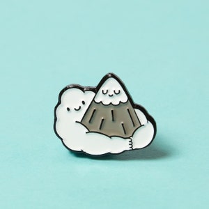May include: A white cloud with a friendly face is hugging a grey mountain with a smiling face. The enamel pin is black and white with a grey mountain.