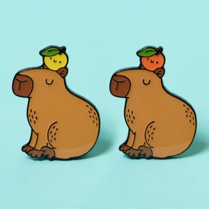 Peut inclure: Deux épingles en émail représentant un capybara brun avec un visage souriant. Un capybara porte un fruit jaune avec une feuille verte sur la tête. L'autre capybara porte un fruit orange avec une feuille verte sur la tête.