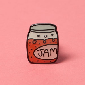 May include: A red and white enamel pin shaped like a jar of jam with a smiling face and the word "JAM" on the label.