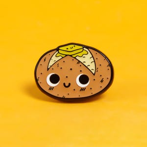 May include: A brown cartoon potato with a smiling face and big eyes. The potato has a pat of butter on top. The potato is an enamel pin.
