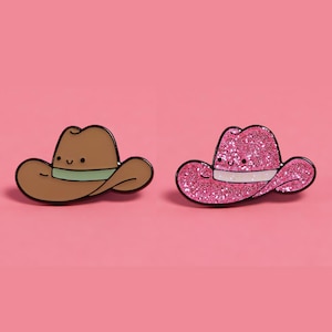 May include: Two cowboy hat enamel pins. One is brown with a green band, the other is pink with glitter and a white band. Both have smiling faces and black outlines. Set against a pink background.