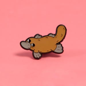 May include: A brown and black enamel pin of a cartoon platypus with a wide smile and big eyes. The platypus is swimming with its tail curled up.