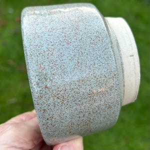 May include: A ceramic container with a speckled, light blue exterior and a cream-coloured rim. The container has a rounded shape and a textured surface, with small brown speckles throughout the blue glaze.