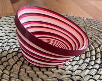 Könnte beinhalten: Eine dekorative Schale mit Spiralmuster. Die Schale hat abwechselnde Bänder in Weiß, Rosa und Burgunder. Sie steht auf einem gewebten, kreisförmigen Platzset. Der Rand der Schale ist einfarbig burgunderfarben.