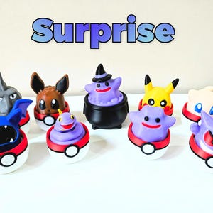 Può includere: Una collezione di personaggi Pokémon colorati, ognuno dei quali emerge da una Poké Ball rossa e bianca. La parola "Sorpresa" è scritta in blu sopra le figure. Le figure includono Pikachu, Eevee e altri.