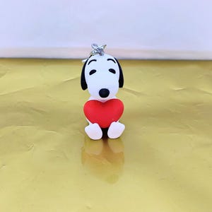 Può includere: Una piccola statuetta di cane bianco con orecchie e occhi neri, che tiene un cuore rosso brillante tra le zampe anteriori. Il personaggio ha un muso arrotondato e un naso nero, con un anello in metallo argentato fissato sulla testa.
