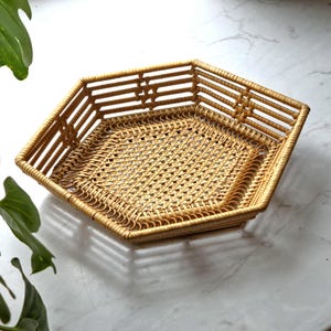 May include: A hexagonal woven rattan tray in a natural tan color. The tray features a woven base and a decorative border. The sides are constructed with horizontal rattan strips. The tray is designed for serving or display.