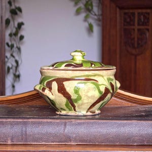 May include: A ceramic lidded bowl with a light green and brown camouflage pattern. The bowl has two small handles and a rounded lid with a small knob. The bowl is sitting on a wooden surface.