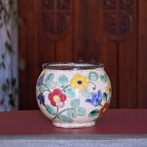 Könnte beinhalten: Kleiner, runder Keramiktopf mit cremefarbenem Boden. Verziert mit bunten Blumenmustern in Rot, Gelb, Blau und Orange, mit grünen Blättern. Der Rand ist dunkelbraun. Der Topf steht auf einer roten Oberfläche.