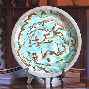 Könnte beinhalten: Eine Keramikschale mit einem einzigartigen Marmordesign. Die Schale hat ein hellblaues und braunes Wirbelmuster mit einem beigen Rand. Sie wird auf einem dekorativen Metallständer präsentiert, der ihr künstlerisches Design hervorhebt. Diese handgefertigte Schale ist ein dekoratives Stück.