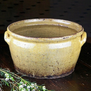 May include: A round, yellow-glazed ceramic pot with two handles. The pot has a wide opening and a slightly textured surface. The bottom edge has a dark brown color. A sprig of white flowers rests on the dark wooden surface.