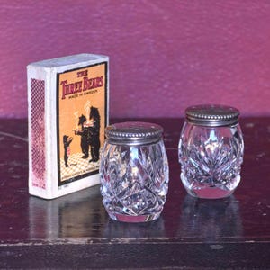 Pode incluir: Dois saleiros transparentes de cristal lapidado com tampas prateadas, ao lado de uma caixa de fósforos. A caixa de fósforos tem um fundo castanho com as palavras "The Three Bears" e uma ilustração de ursos. Fabricado na Suécia.