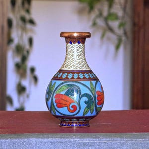 Könnte beinhalten: Eine kleine, dekorative Vase mit kupferfarbenem Rand und Boden. Die Vase hat einen hellblauen Körper mit floralen Designs in Rot, Grün und Blau. Der Hals hat ein weißes und goldenes Muster. Ein schönes Stück für die Inneneinrichtung.