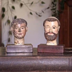 Könnte beinhalten: Zwei antike, handbemalte Holzbüsten männlicher Figuren. Eine Büste hat kurzes Haar, die andere einen Bart und eine Kopfbedeckung. Beide sind auf rechteckigen Holzsockeln montiert. Die Büsten sind in gedämpften Tönen bemalt und weisen sichtbare Alters- und Gebrauchsspuren auf.