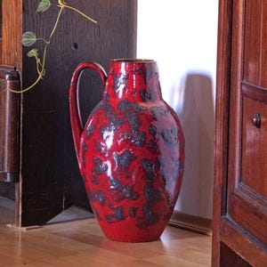 Könnte beinhalten: Ein großer, roter Keramikkrug mit Henkel und dunklem, gesprenkeltem Muster. Die Vase steht auf einem Holzboden, mit einer dunklen Holztür und einer weißen Wand im Hintergrund. Der Krug ist etwa 46 cm hoch.