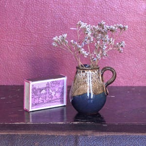 Könnte beinhalten: Eine kleine Keramikvase mit Henkel, gefüllt mit Trockenblumen. Die Vase ist braun und blau. Eine Streichholzschachtel mit einer lila-weißen Illustration eines Gebäudes befindet sich neben der Vase.