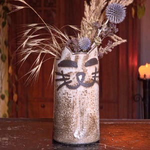 Puede incluir: Un jarrón de cerámica con diseño de cara de gato, lleno de flores y hierbas secas. El jarrón es de color blanco roto con detalles en negro, y las flores incluyen cardos morados y hierbas de color beige. El jarrón mide unos 20 cm de alto.