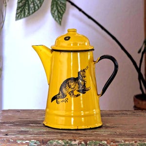 以下が含まれることがあります： 黒いハンドルと注ぎ口が付いた、鮮やかな黄色のエナメルコーヒーポット。ポットには、猫のような動物の黒いイラストが描かれています。ポットには、上部に小さな黒い円が付いた蓋が付いています。ポットは木製の表面にあります。
