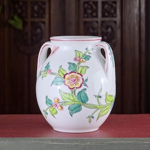 May include: A white ceramic vase with two handles and floral designs. The vase features a pink rim and handles, with green leaves and yellow, pink, and red flowers. The vase is on a red surface.