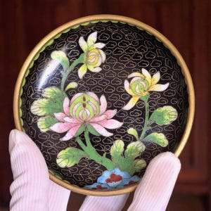 Pode incluir: Um pequeno prato redondo decorativo com fundo preto e um desenho floral. As flores são rosa, amarelas e brancas, com folhas e caules verdes. O prato tem uma borda dourada.