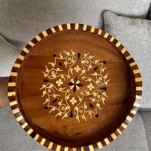 Peut inclure: Un plateau rond en bois présentant un motif central incrusté de volutes claires et de formes en losange sombres. Le bord du plateau est orné d'un motif alterné de segments de bois clair et foncé disposés en une bordure rayée.