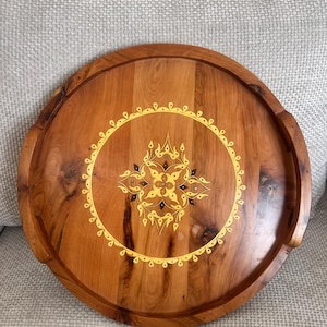 Peut inclure: Un plateau de service rond en bois avec une finition polie, mettant en valeur un grain de bois naturel. Le centre est orné d'un motif floral peint en jaune à l'intérieur d'une bordure circulaire, et le plateau possède deux poignées intégrées.