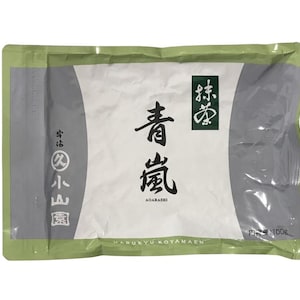 May include: A sealed package of Japanese matcha powder. The package has a white central area with a green border and grey accents. The text "青嵐 AGARASHI" is in black, with Japanese characters and a "100g" label.