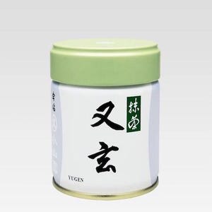 Puede incluir: Un recipiente cilíndrico blanco con tapa verde claro. El recipiente tiene caracteres japoneses y la palabra "Yugen". Probablemente sea para té o un producto similar.