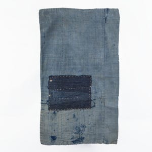 May include: A rectangular piece of faded blue fabric featuring a fine grid pattern. A dark navy blue rectangular patch is stitched onto the center with visible white thread. The fabric shows signs of wear with scattered dark blue spots and frayed edges.