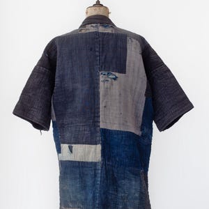 May include: A short-sleeved jacket constructed from various rectangular patches of indigo-dyed fabric in shades of dark navy, slate blue, and faded grey. The garment features visible vertical stitching throughout and displays signs of wear with small frayed areas.