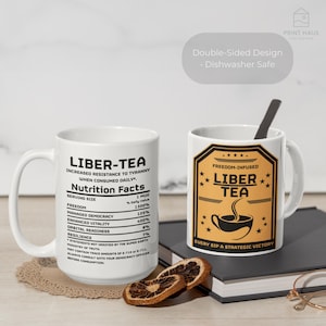 May include: Two white ceramic mugs with black text and graphics. One mug features "LIBER-TEA" and a nutrition facts label. The other mug has a gold and black label that says "LIBER TEA" with a cup of tea graphic. A spoon rests in the second mug.