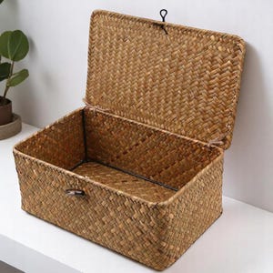 May include: A rectangular woven storage basket with a hinged lid, made from natural-coloured seagrass. The lid is partially open, revealing the interior. The basket has a metal frame and a small latch. Ideal for home organisation.