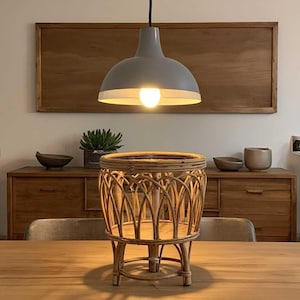 Könnte beinhalten: Eine graue Pendelleuchte beleuchtet einen Pflanzenständer aus natürlichem Rattan. Der runde Pflanzenständer hat ein gewebtes Design und drei kurze Beine. Die Szene spielt in einem Esszimmer mit Holzmöbeln.