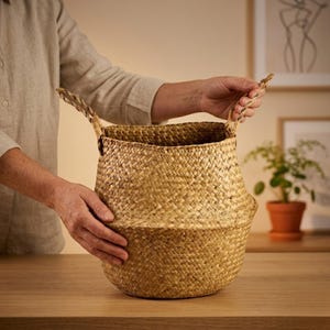 May include: A woven basket with two handles is held by a person. The basket is made of a light brown material and has a textured appearance. The basket is sitting on a wooden surface.