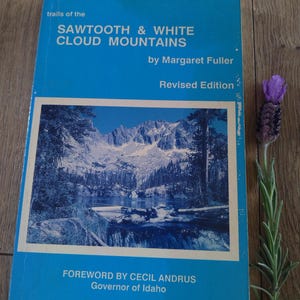 May include: A blue book titled "Trails of the Sawtooth & White Cloud Mountains" by Margaret Fuller. The cover features a mountain landscape image. A sprig of lavender is next to the book.