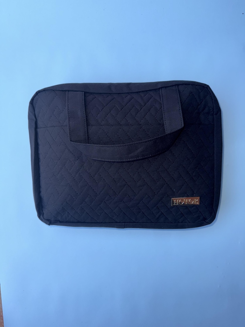 May include: Black quilted laptop sleeve with a handle. The sleeve has a geometric pattern and a gold-colored rectangular label that reads "HOTOR". The background is a light blue.