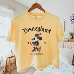 Puede incluir: Camiseta amarilla mantequilla con el logotipo de Disneyland y un gr&aacute;fico de Minnie Mouse. El gr&aacute;fico incluye el texto "Est 1955" y "California". La camiseta est&aacute; colgada en una percha de madera.