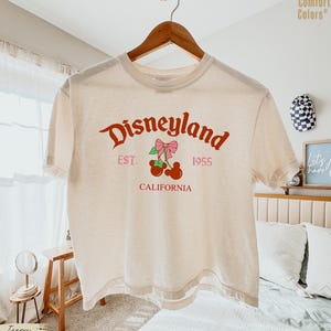 May include: Ivory-colored t-shirt with the word "Disneyland" in a red script font. Below the word is "EST. 1955" and "CALIFORNIA" in a pink font. A graphic of cherries with a pink bow is in the center.