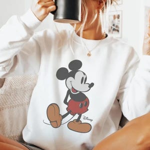 Puede incluir: Sudadera blanca con un gr&aacute;fico de Mickey Mouse. El personaje de dibujos animados est&aacute; en negro, rojo y marr&oacute;n. El logotipo de Disney est&aacute; impreso debajo. La sudadera es de manga larga y tiene cuello redondo. Se sostiene una taza negra.