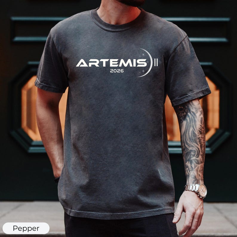 May include: A dark gray t-shirt with the text "ARTEMIS II 2026" in white. The design includes a crescent moon and stars. The shirt is a short-sleeved crew neck.